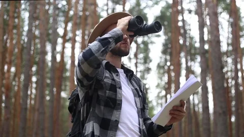 Man in a hat holds a map in and looks through binoculars while hiking Stock-Footage 149946617