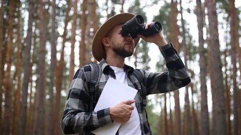 Man in a hat holds a map in and looks through binoculars while hiking Stock Footage 149946623