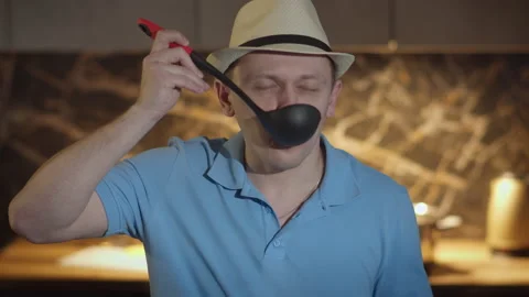 A man in a hat with a ladle stands in front of the kitchen, tasting food from 스톡 동영상 165254100
