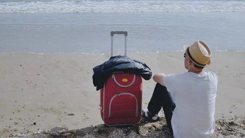 Man in hat with red suitcase sitting on the seashore, vacation Stock Footage 155941557