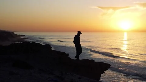Man in hat on rocky beach looking beautiful sunset sky ocean landscape nature Stock Footage 149359025