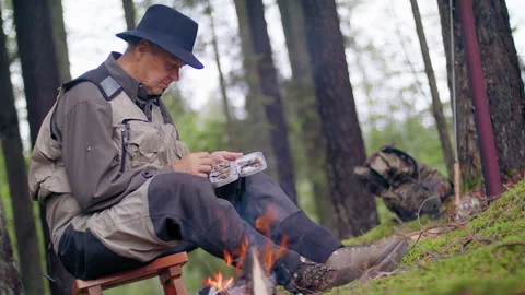 Man in hat sits against the background of fishing gear, fire  and forest Video stock 141254810