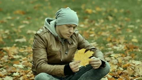 Man in a hat sits among the fallen leaves, looking at a yellowed maple leaf, the Stock Footage 165901370