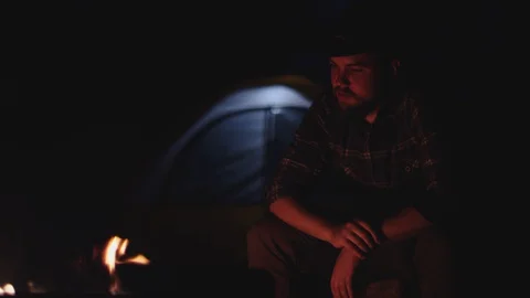 Man in hat sits by a campfire at night in slow motion Video stock 128779989
