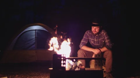 Man in hat sits by a campfire at night in slow motion Stock-Footage 128780297