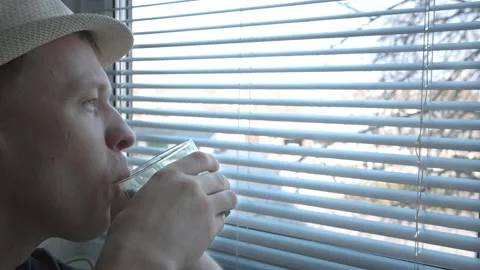 A man in a hat sits near the window, drinks a drink and looks with interest Stock Footage 156301238