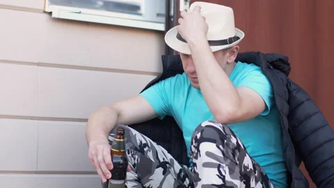 Man in a hat sits in thought with a bottle of beer on the doorstep of the house Stock Footage 153859700
