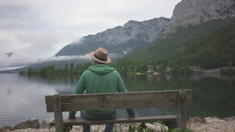Man in hat sitting and looking at view of mountain lake Grundlsee in fog Stock Footage 132060145