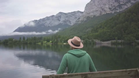 Man in hat sitting and looking at view of mountain lake Grundlsee in fog Stock Footage 132060487