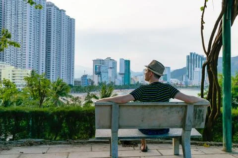 Man in hat is sitting on bench looking at beach with modern buildings Stock Photos