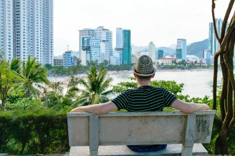 Man in hat is sitting on bench looking at beach with skyscrapers Stock Photos