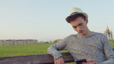 Man in hat sitting on bench with mobile phone in hand, using mobile app 스톡 동영상 304272224