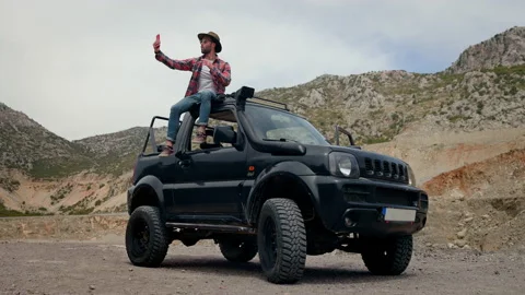 Man In Hat Sitting On Car. Stylish Young Guy In Middle Of Rocky Terrain. Stock Footage 155803336