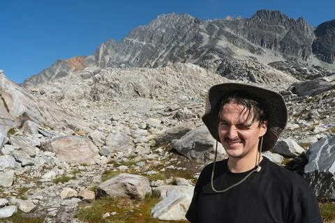 A man in a hat smiles in front of a mountain landscape with rocks and sky. Stock Photos