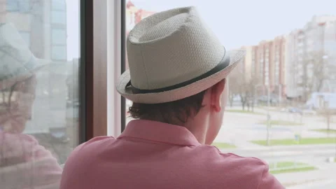 Man in a hat standing in the window opening looks at the street and drinks a Stock Footage 158380936