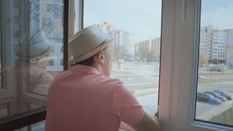 A man in a hat stands on the balcony looking at the street through an open Stock Footage 165251094