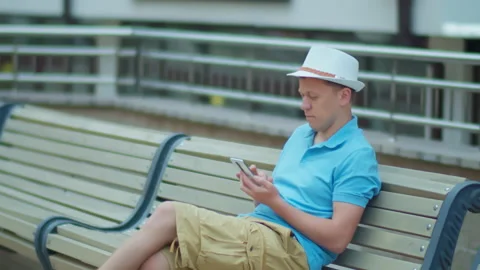Man in hat uses mobile phone while sitting on city bench, camera tracking Stock Footage 148228033