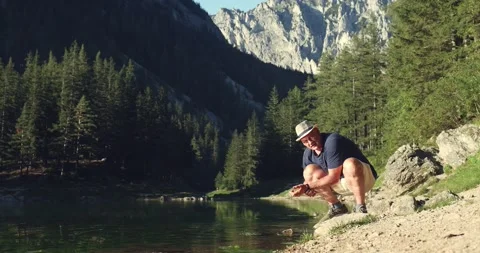 A man in a hat washes his hands in a mountain lake in the Alps Video stock 246491851