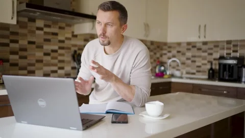 Man Having Video Call and Gesturing at Laptop in Modern Kitchen. A man sitt.. Stock Footage 331971360