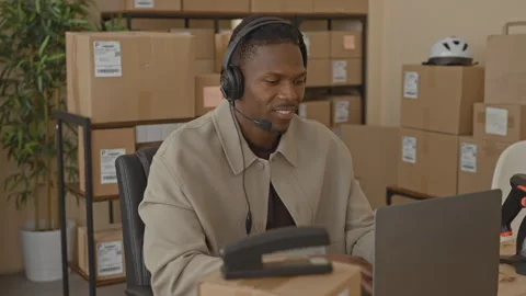 Man with headset smiles while typing on laptop and handling parcels at desk.. Stock Footage 337434737