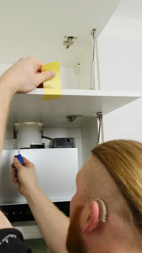 Man with hearing aid installs hinges on cabinet door in kitchen Stock Footage 306458719