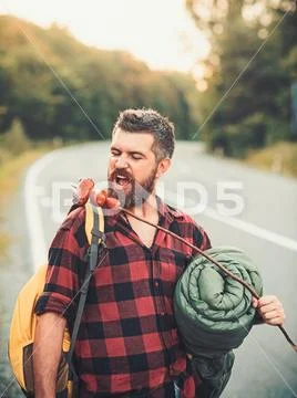 Man hiker eat sausage on stick. Bearded man with food hiking on road. Hipster Stock Photos