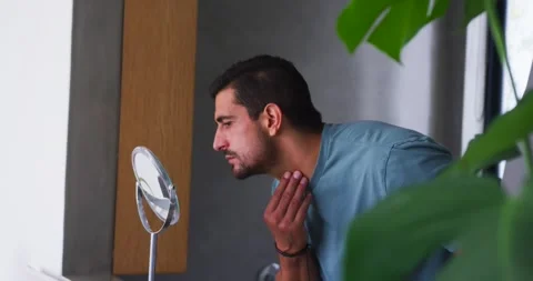 Man in his 30s approaching tabletop mirror at bathroom vanity, inspecting Stock Footage 344949578
