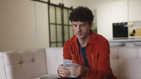 Man in his 30s counting dollar bills on sofa, happy with investment gains Stock Footage 337908470