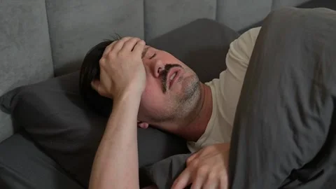 A man in his 30s is lying in bed, showing signs of discomfort from a headache. 스톡 동영상 344527285