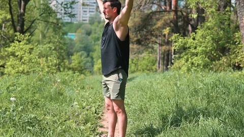 A man in his 30s performs stretching exercises in a green outdoor area with Stockbeeldmateriaal 339016202