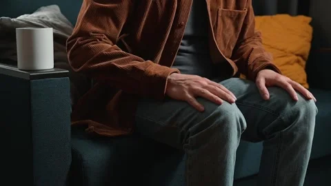 A man in his 30s is seated on a couch, gently tapping his knees while holding a Stock Footage 339097556