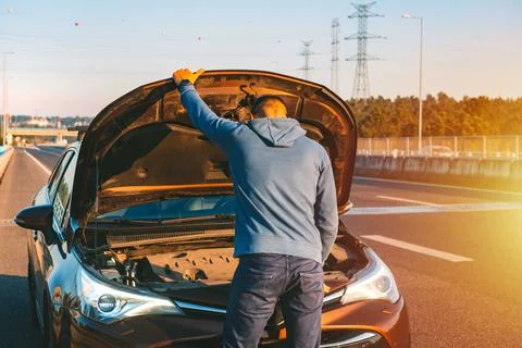 Man with his broken car checking engine on the highway roadside at sunset Stock Photos