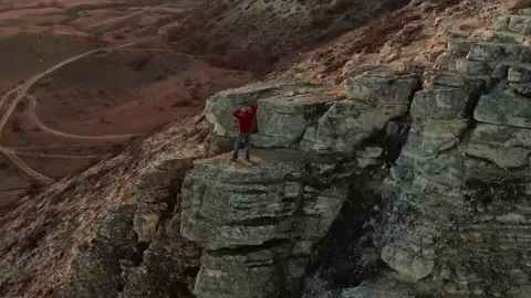 A man with his hands folded behind his head stands on a ledge of a rock and watc Stock Footage 150566507