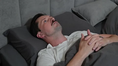 A man in his thirties is lying in bed with his hands clasped, looking Видео 344527257
