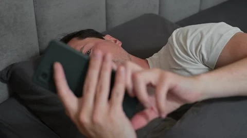 A man in his thirties is waking up in bed, looking tired and checking his phone. 動画素材 344527297
