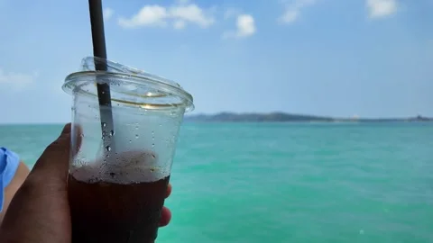 Man holdign iced coffee in plastic cup against sea scape background. Stockbeeldmateriaal 274190014