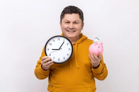 Man holding big wall clock and piggy bank with banknote inside, looking smiling Stock Photos