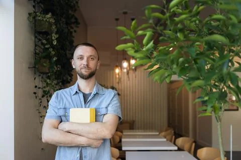 Man holding books while standing in a cafe with plants, working on online e.. Fotos de archivo