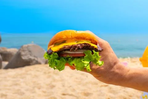 Man holding burger in a hand against beach background Stock Photos
