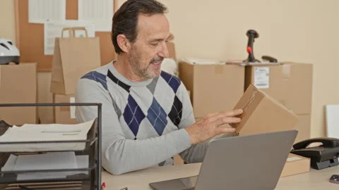 Man holding a cardboard package and inspecting label at a desk with laptop .. Stockbeeldmateriaal 337443868
