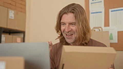 Man holding cardboard package smiling at laptop amid stacked boxes and ship.. Stock Footage 337375904