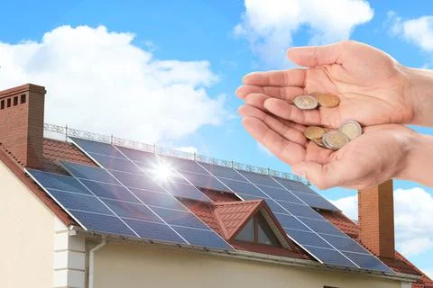 Man holding coins against house with installed solar panels. Renewable energy Stock Photos