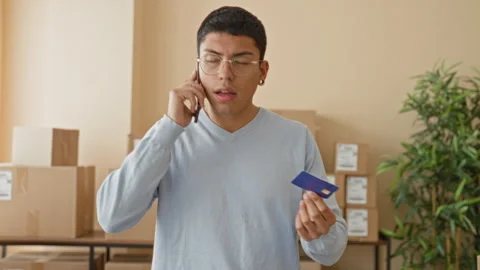 Man holding creditcard while talking on smartphone in a warehouse packing a.. Stock Footage 339252692