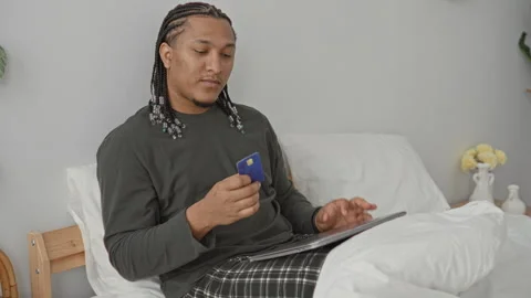 Man holding a creditcard while using a laptop on a bed in a bedroom, braids.. Stock Footage 347813725