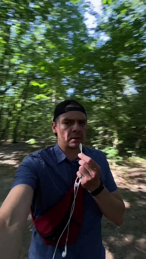 Man holding earbud microphone while walking through sunlit deciduous forest Видео 344584594