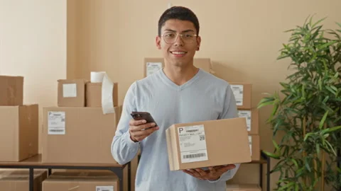 Man holding parcel and smartphone, smiling while checking a shipping label .. Video stock 337431583