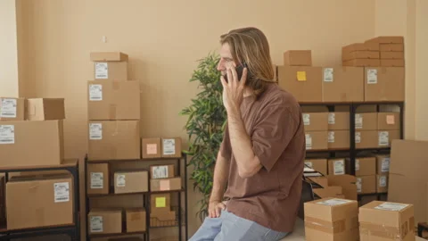 Man holding smartphone to ear with hand, surrounded by stacked packages in .. Stock Footage 339189653