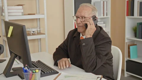 Man holding smartphone to ear while seated at a desk with desktop computer,.. Stock Footage 339198945