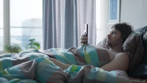 Man holding smartphone lying in bed in the morning, shoked, surprised news Stock Footage 277429615