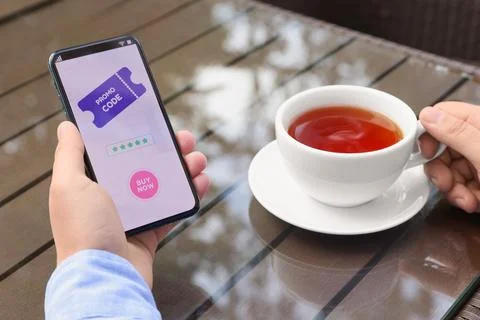 Man holding smartphone with promo code and cup of tea at wooden table outdo.. Stock Photos